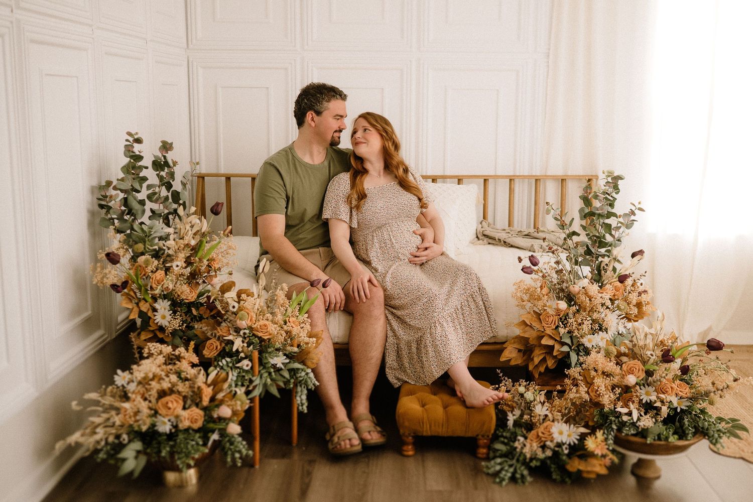 A couple sits on a wooden bench surrounded by beautiful peach and cream floral arrangements in a light-filled studio setting.