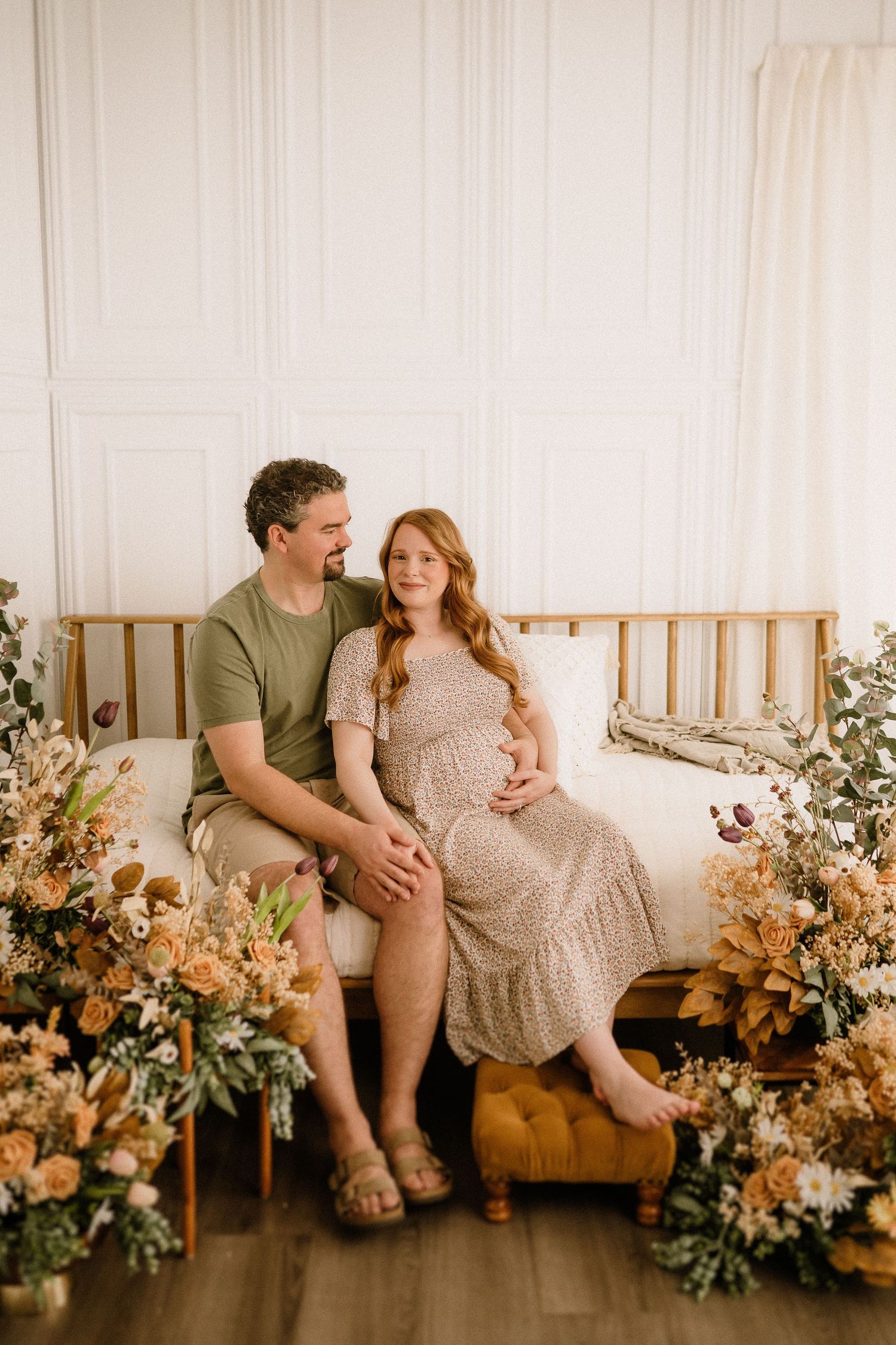 Romantic maternity photo of couple sitting on gold bench surrounded by peach and cream floral arrangements.