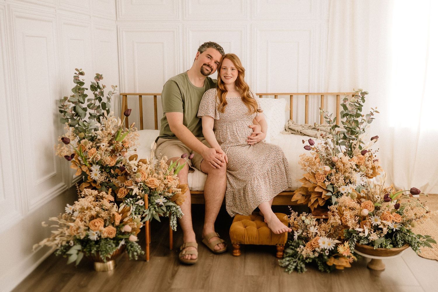 Couple sits on wooden bench surrounded by peach and cream floral arrangements in bright white studio setting.
