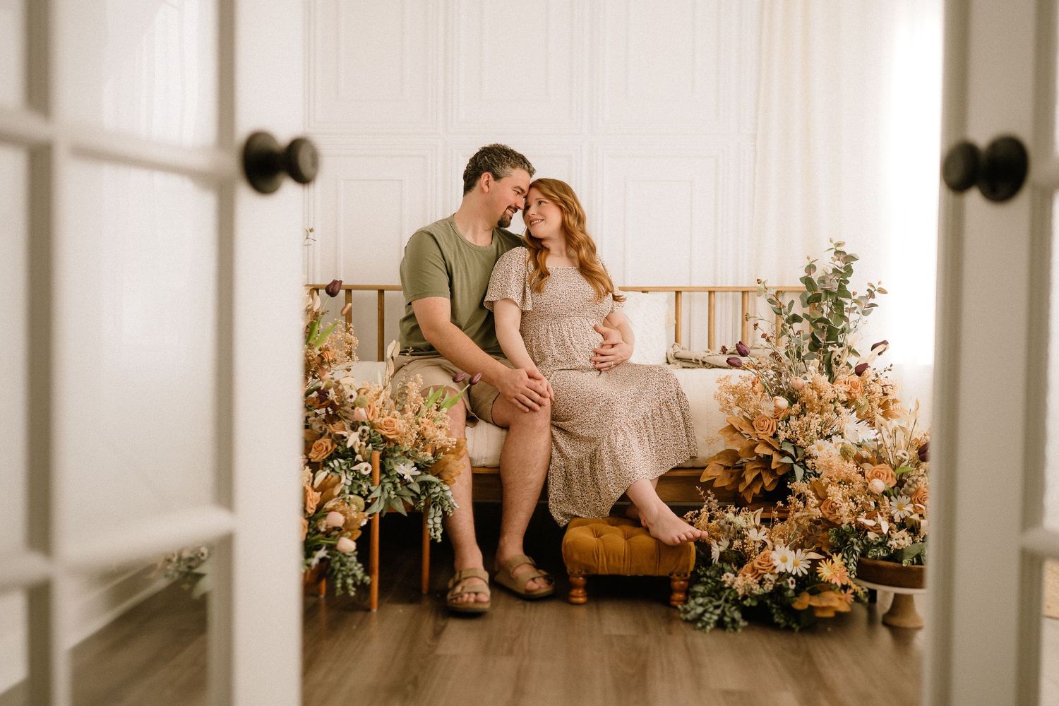 A couple sits together on a wooden bench surrounded by autumn-colored floral arrangements in a bright sunlit room.