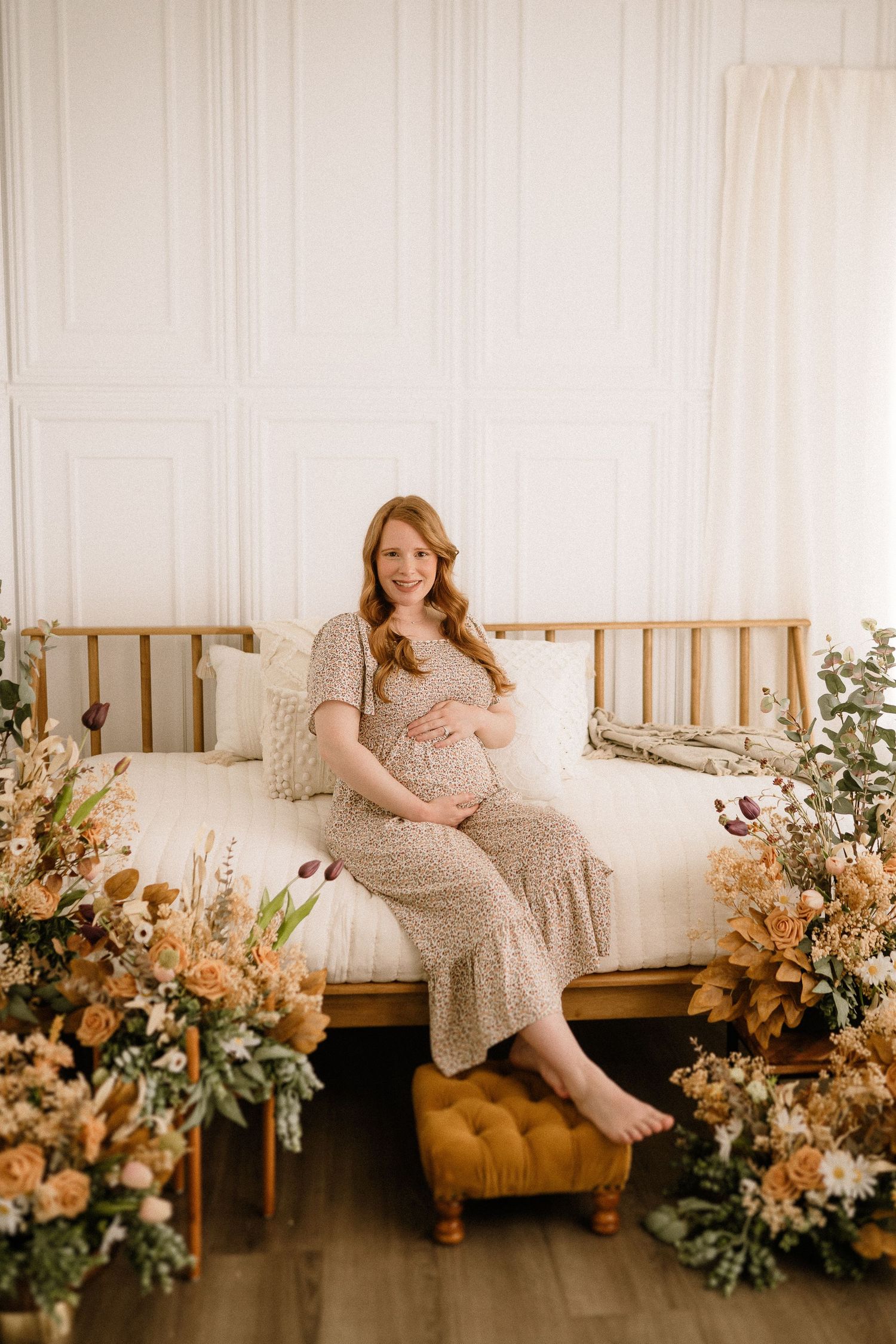 Expectant woman in a beige dress lounges on a daybed surrounded by romantic peach and cream floral arrangements.