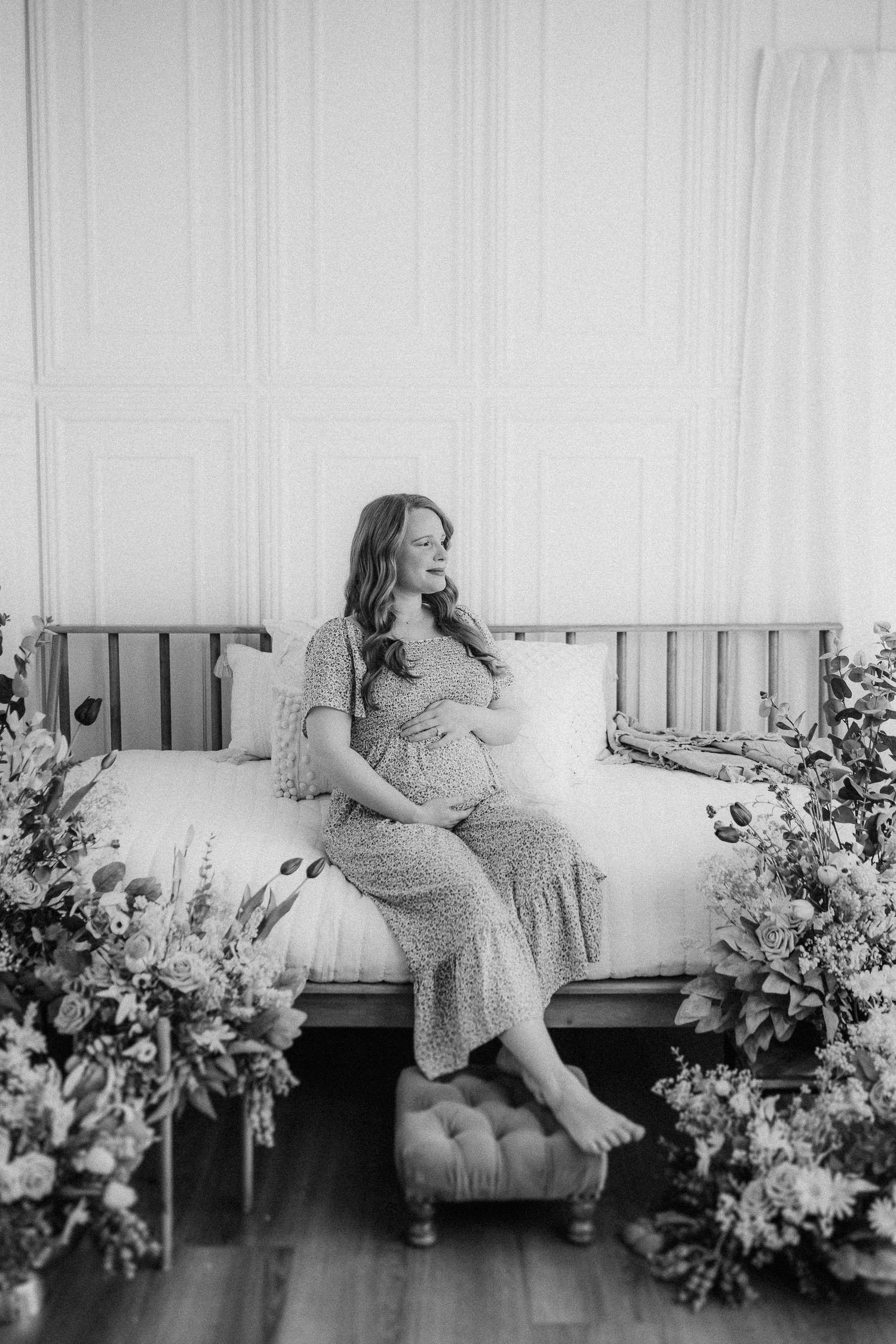 Black and white maternity photo showing an expecting mother in a polka dot dress sitting on a daybed surrounded by flowers.