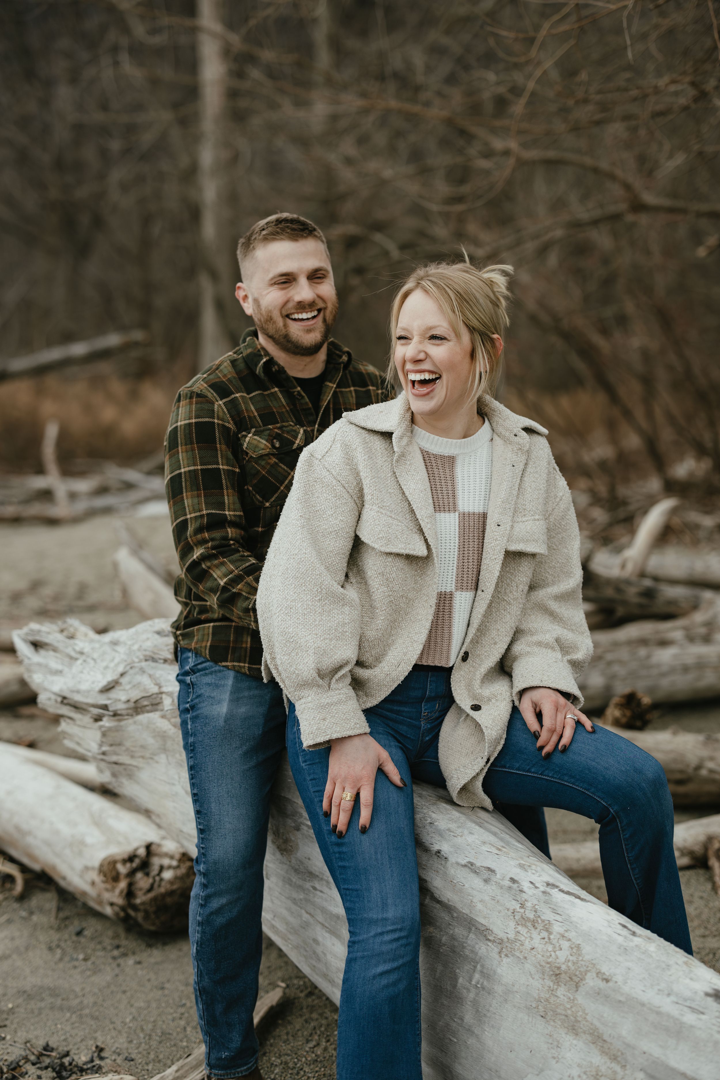 Couple sitting together on driftwood logs at beach sharing candid laughs during casual outdoor photoshoot.