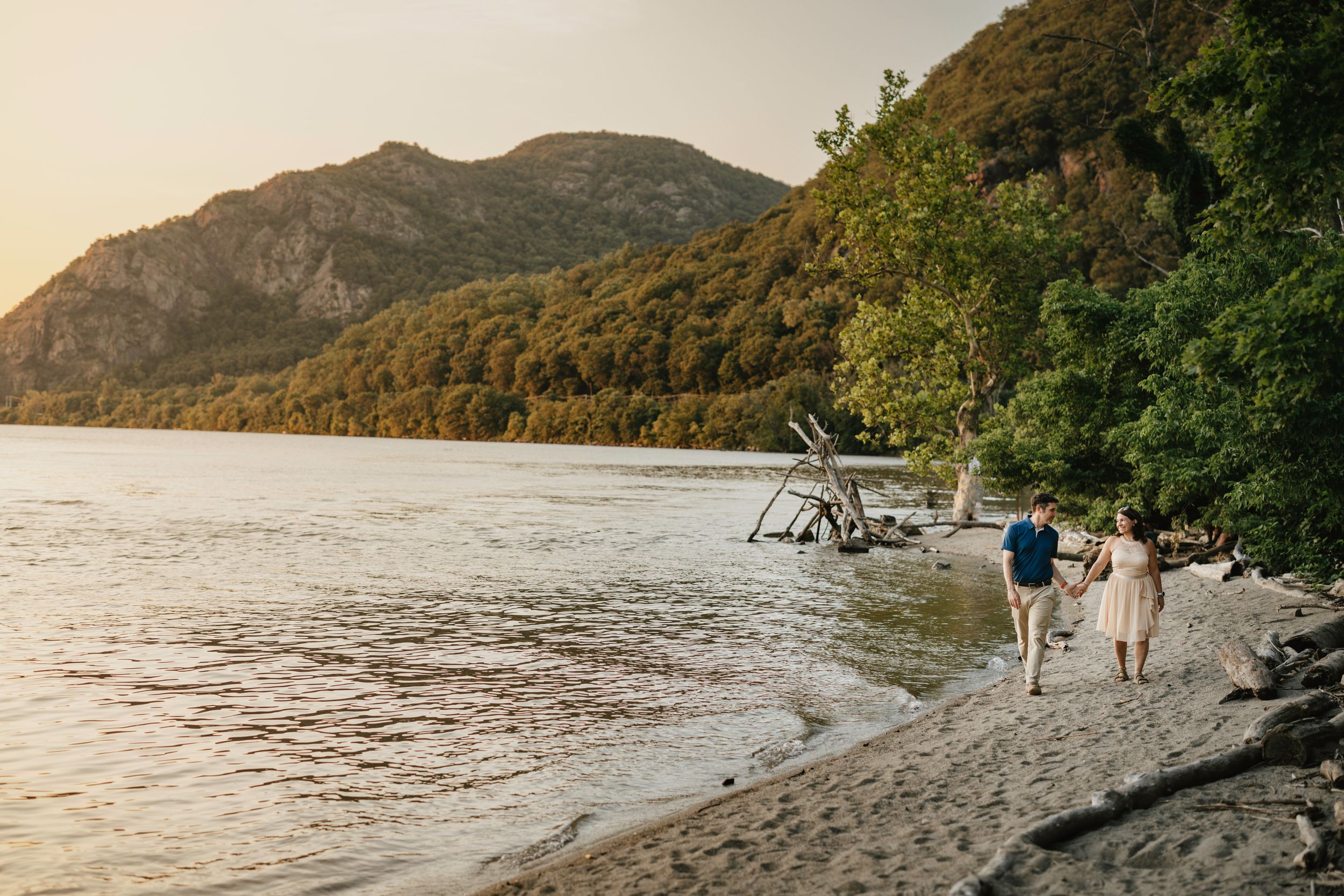 A seaplane rests on a rocky beach at sunset with mountains in the background as two people walk along the shore.