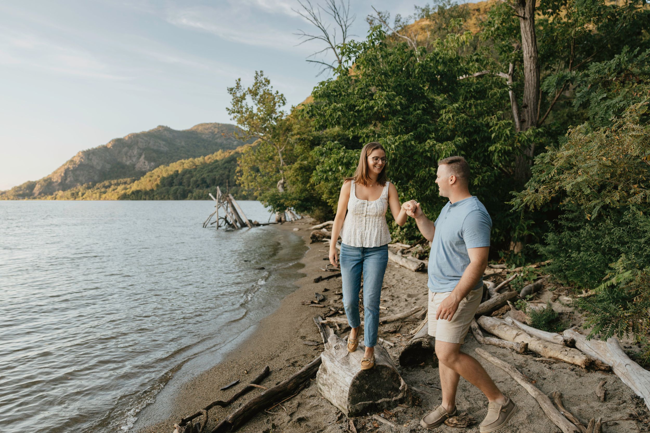A couple walks along a scenic lakeside trail during sunset with mountains and trees in the background.