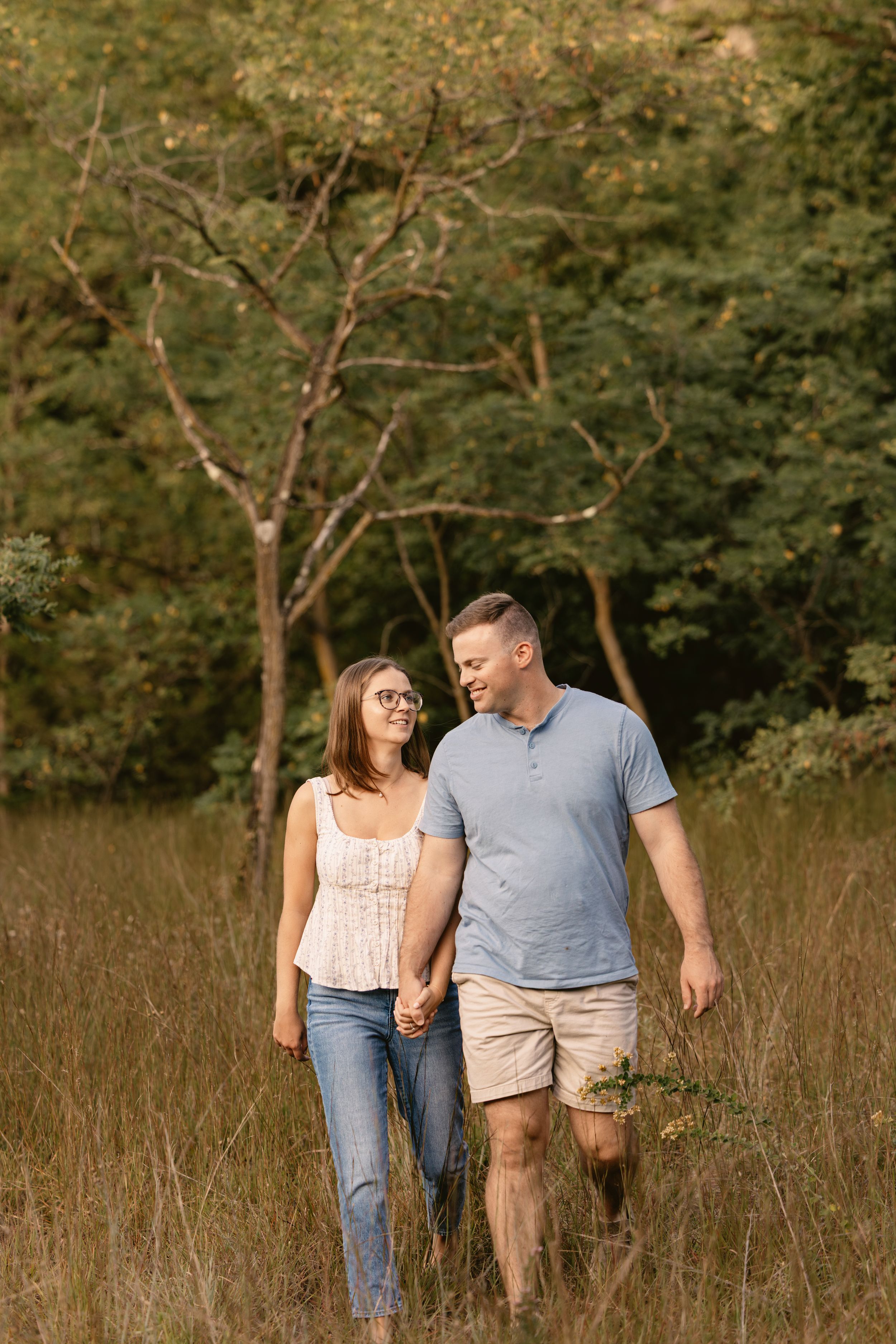 A couple walks hand in hand through a golden meadow at sunset surrounded by trees in the background.