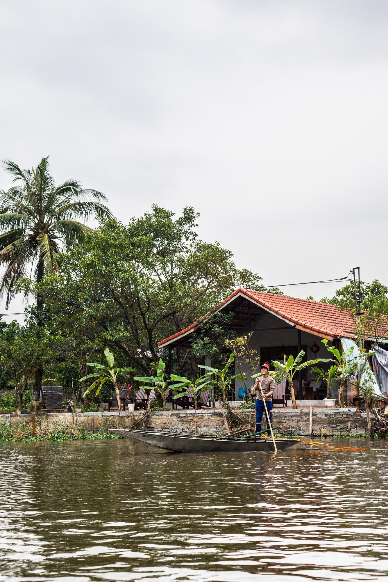 A person poles a wooden boat past traditional stilt houses along a river in Vietnam with palm trees and lush vegetation.