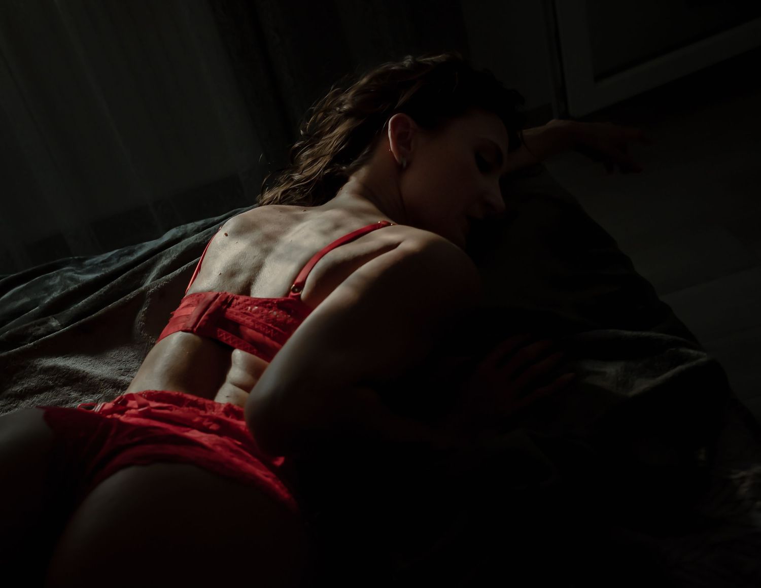 Woman in red lingerie lies dramatically in moody low-key lighting showing back and shoulders.
