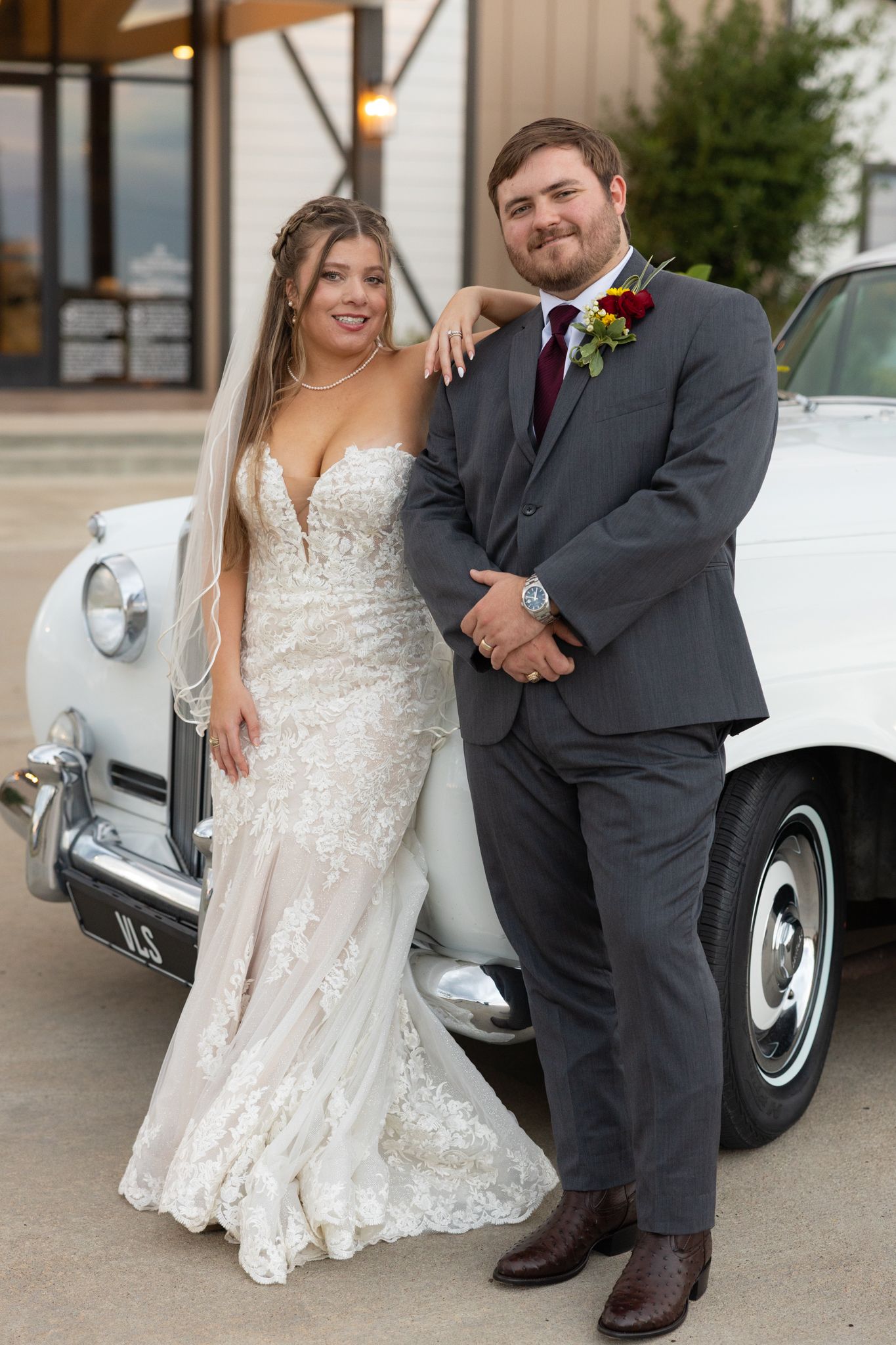 Newlywed couple poses together by a classic vintage car in formal wedding attire.