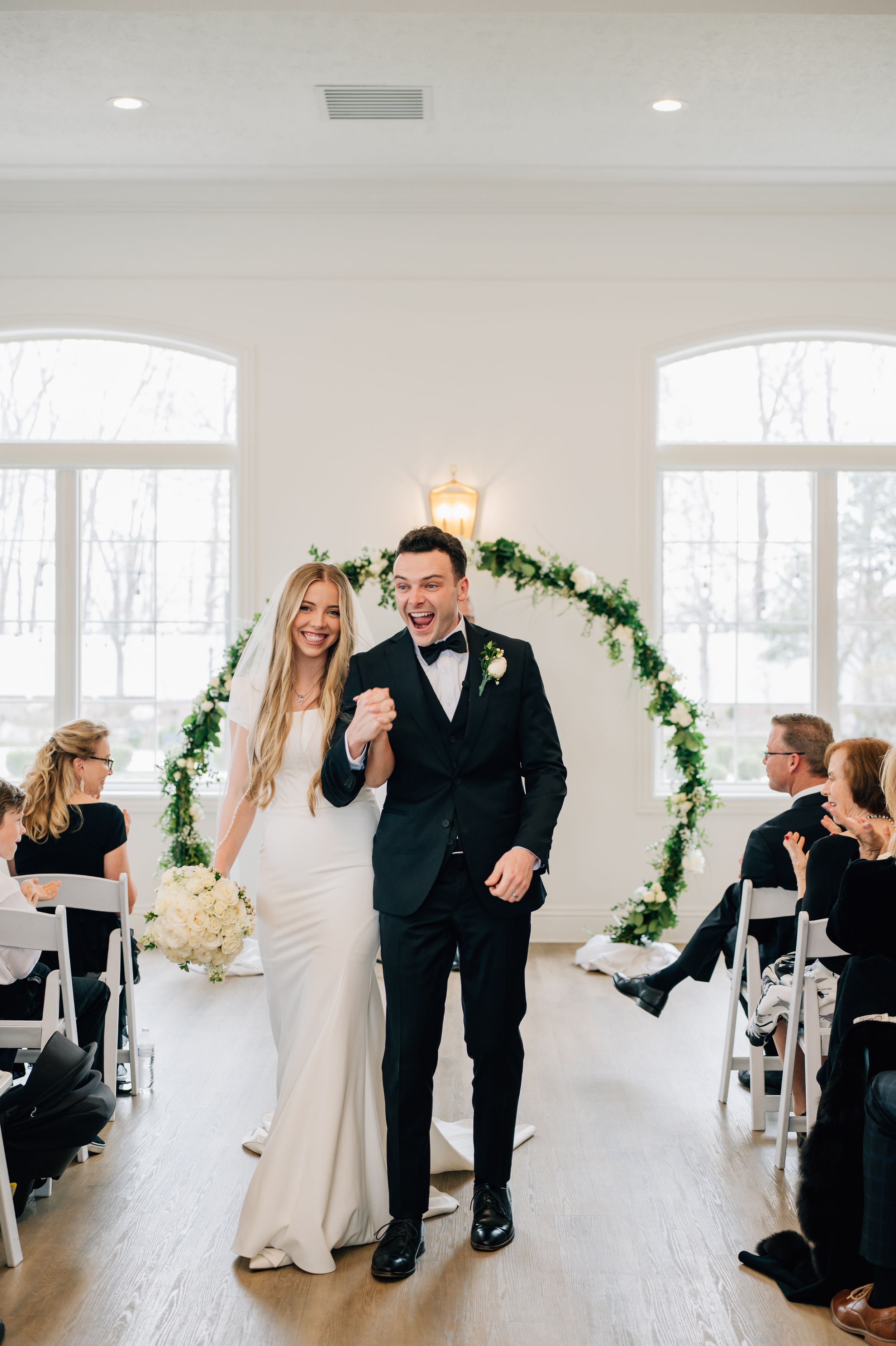 A joyful couple celebrate their wedding ceremony under a floral arch in an elegant white room with large windows.