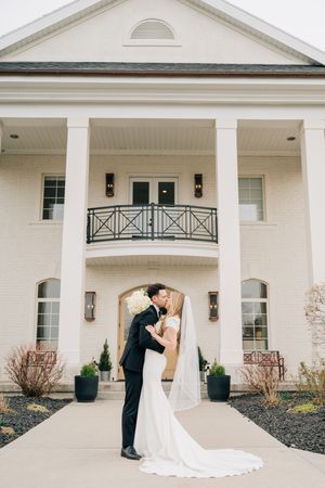 A couple shares an intimate moment outside a grand white mansion with classical columns and elegant architecture.