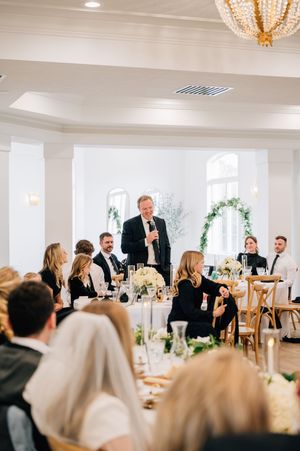 Wedding reception in an elegant white room with chandelier as guests gather at tables while someone gives a speech.