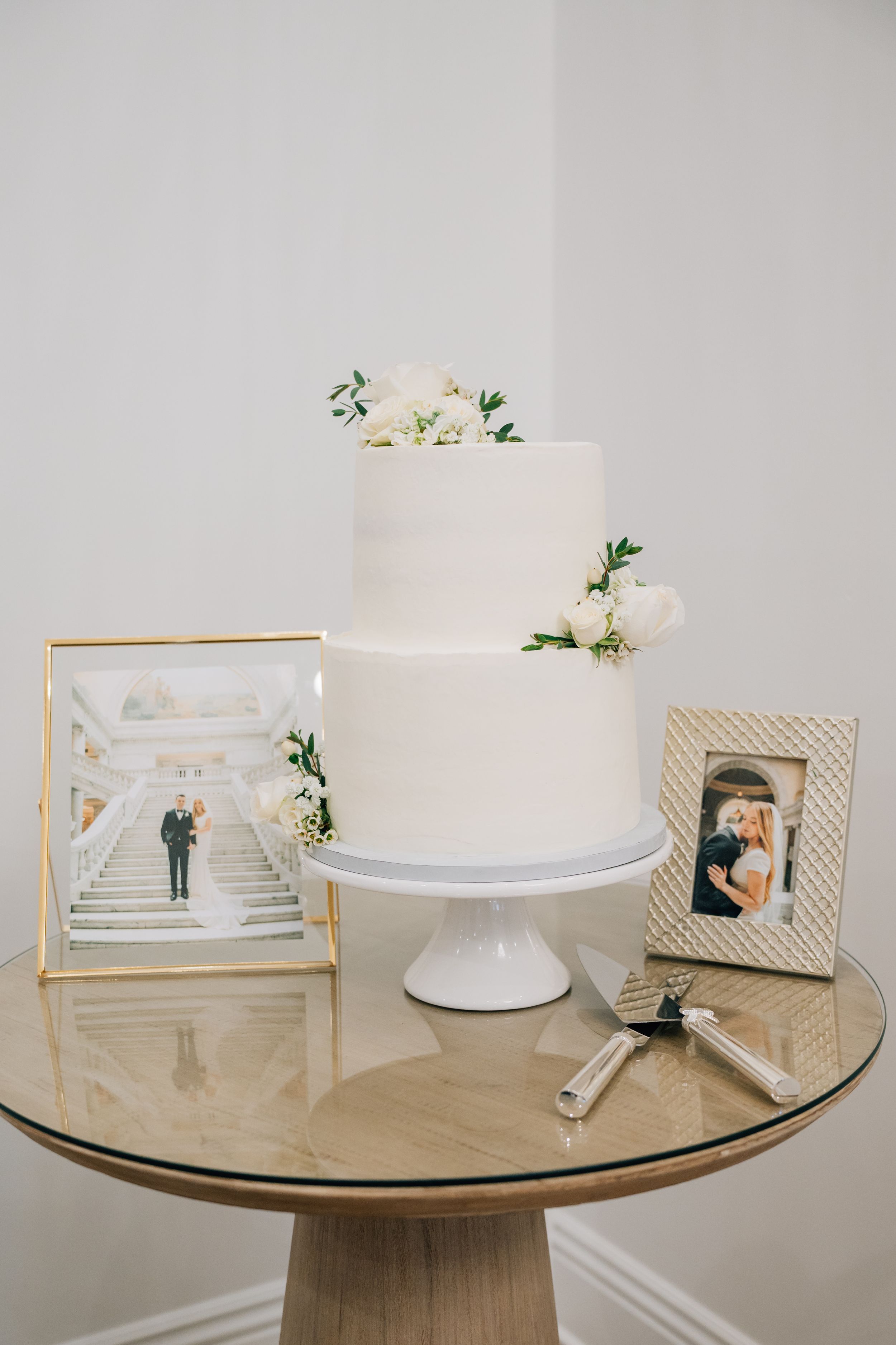Elegant white tiered wedding cake with floral accents displayed on round table next to framed photos and wedding decor.