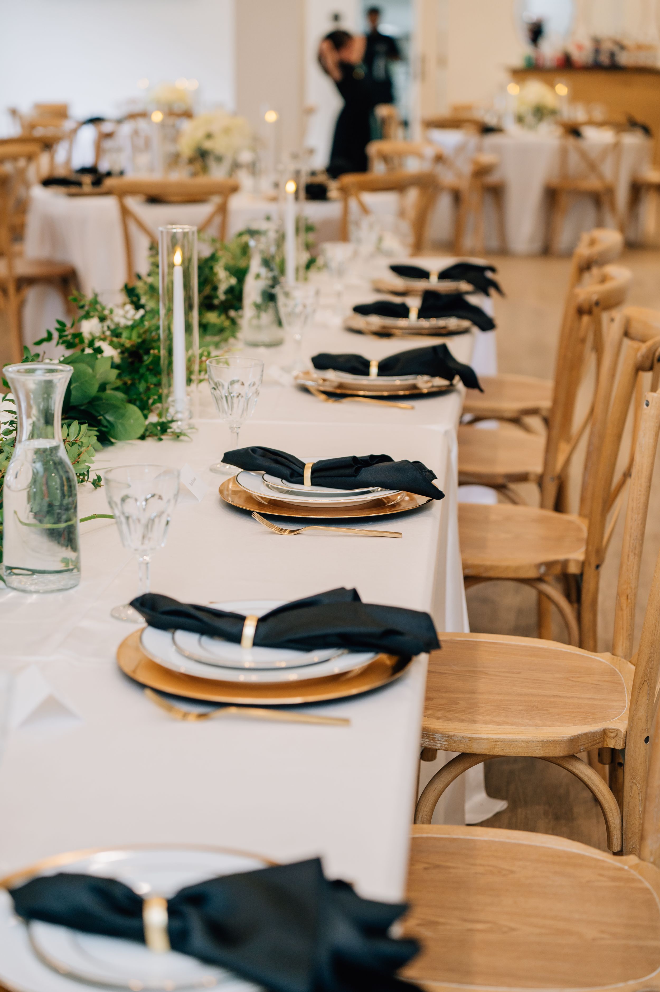 Elegant wedding reception table setting with white linens, rustic wood chairs, and black napkins on gold charger plates.