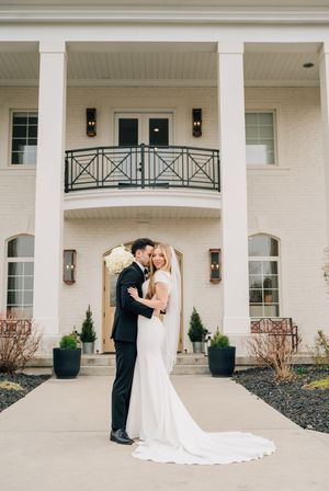 A couple embraces outside a grand white mansion with columns and a balcony, surrounded by elegant landscaping.