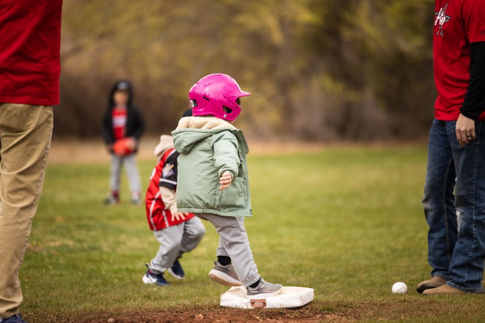 First game of the season little girl jumps to first base at little league game in a jacket and pink helmet. Sports photography.