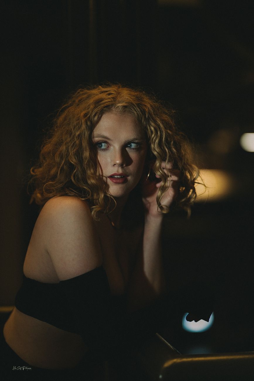 Dramatic moody portrait of a figure with curly red hair in a dark setting with soft ambient lighting.