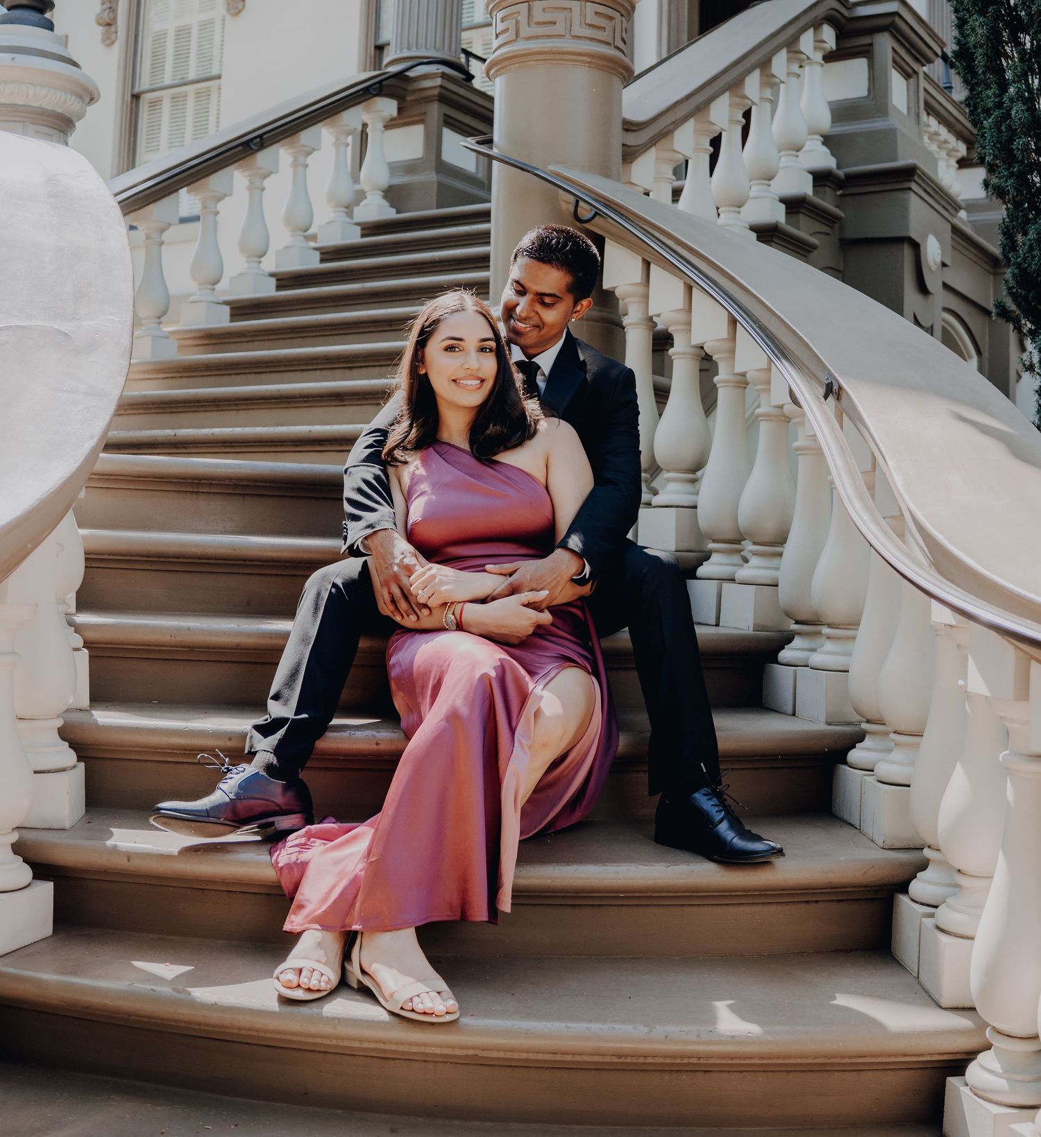 A couple holding hands and standing on elegant stone steps with one wearing a black suit and the other in burgundy attire.