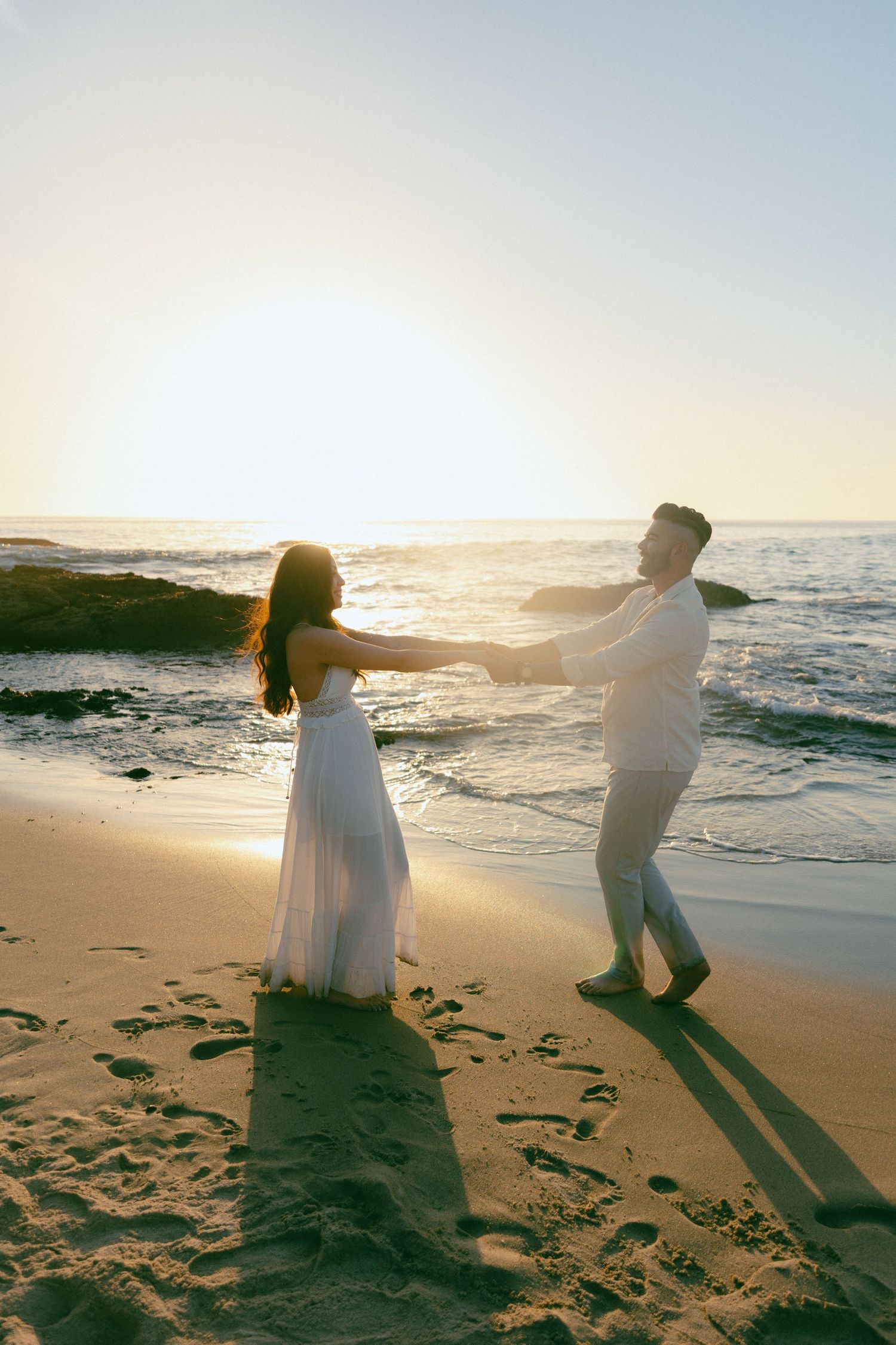 A romantic couple dances together on a beach at sunset with ocean waves and golden light in the background.