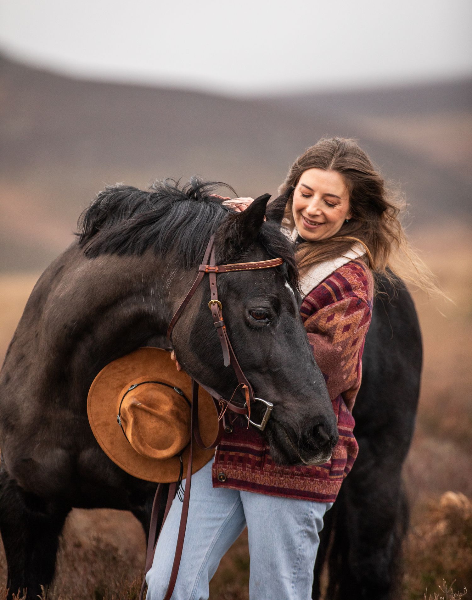 A beautiful white horse stands with its handler in brown winter attire on a hillside with dried vegetation.