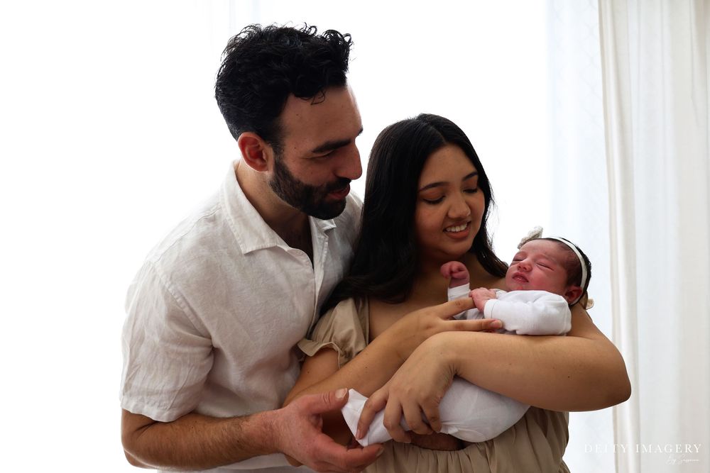 A loving couple shares a joyful moment with their newborn baby wearing white in a bright room by a window.