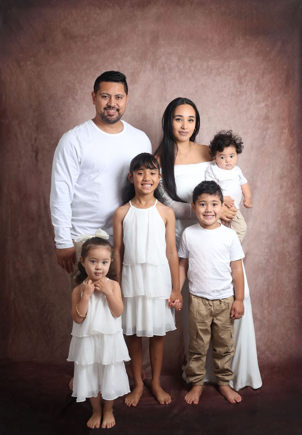 Family portrait series showing different groupings in white and beige outfits against a brown backdrop.
