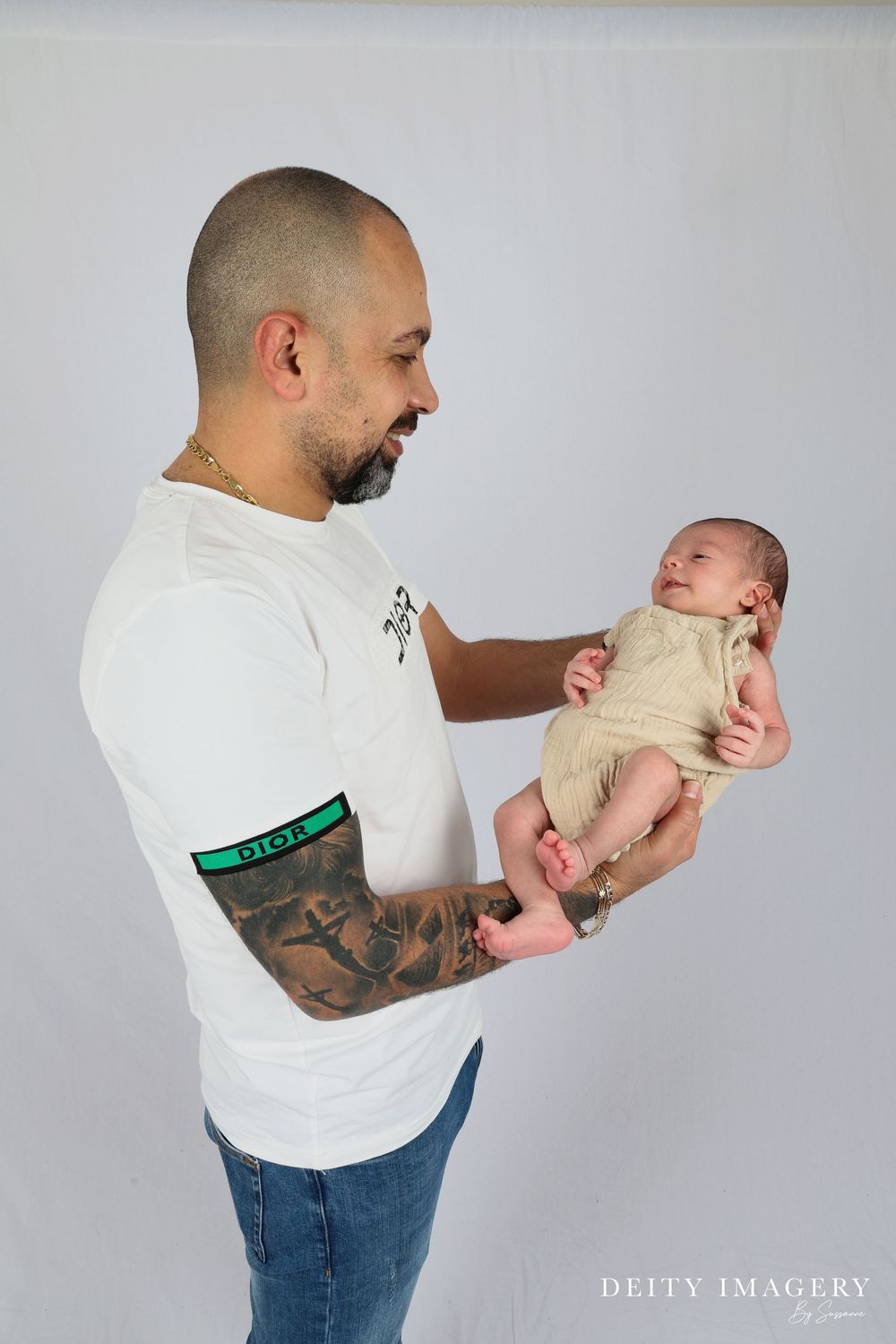 A person in a white t-shirt with a tattooed arm holds a newborn baby against a light gray studio backdrop.