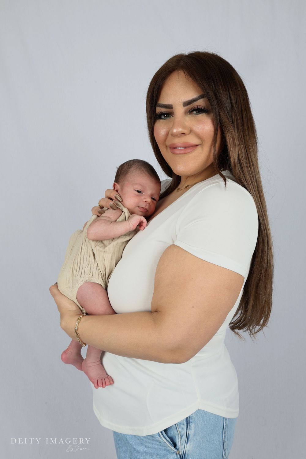 Mother in white shirt holds sleeping newborn baby in tender embrace against gray studio backdrop.