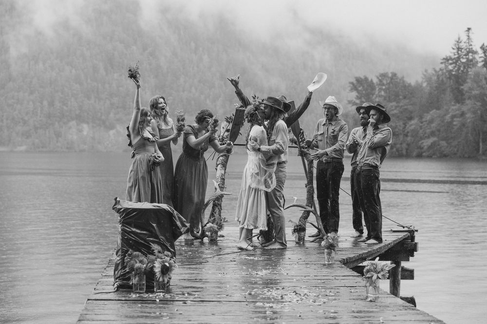 Black and white photo of a celebratory group dancing and posing on a wooden dock over water.