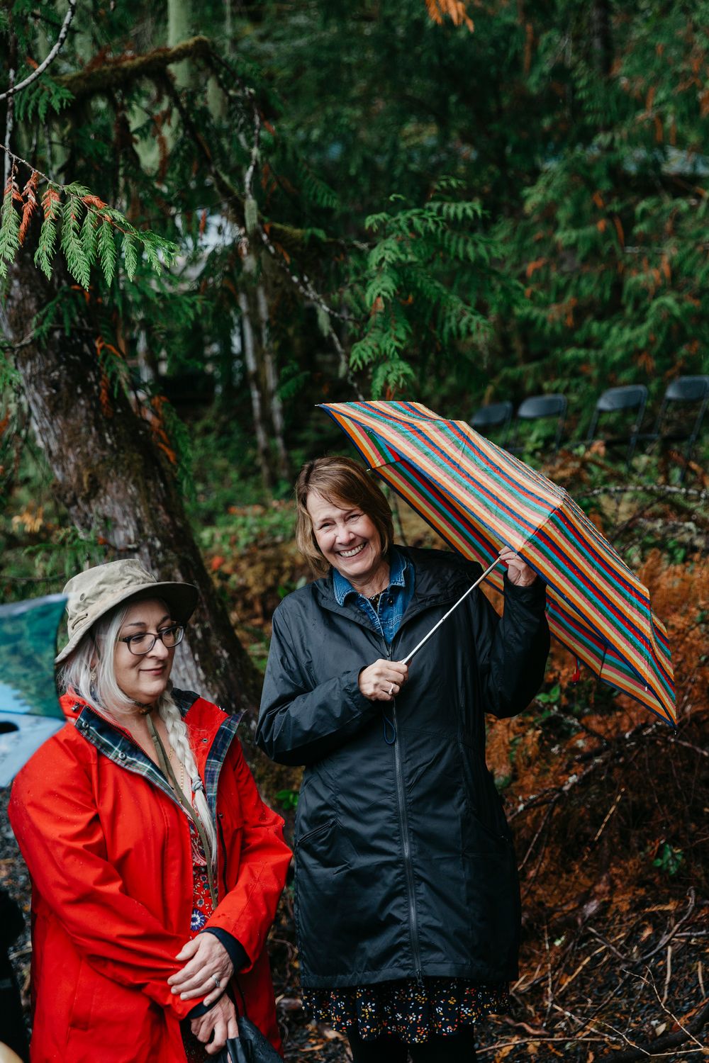 Two people in rain gear smile while holding umbrellas in a lush forest setting.