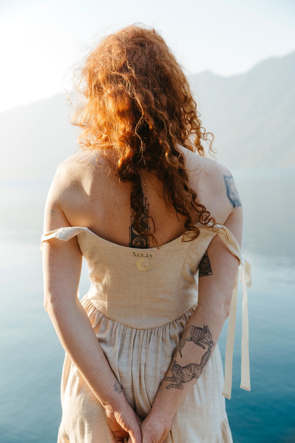 Back view showing intricate tattoos and dress details against serene water landscape.