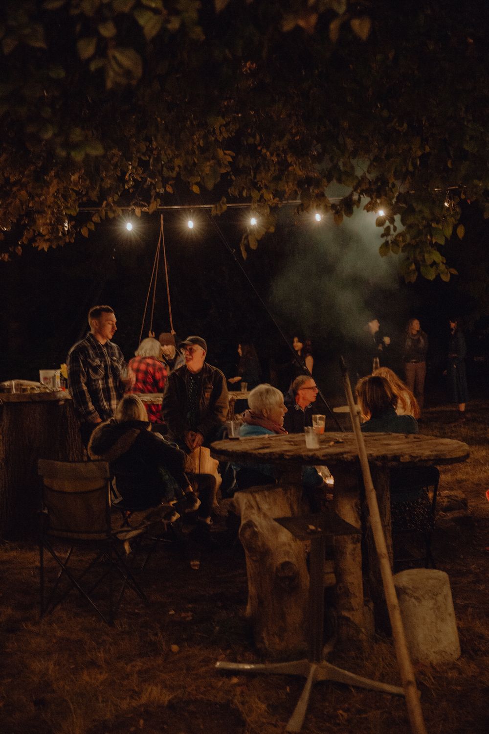 People gathered around wooden tables under string lights at a nighttime outdoor event.