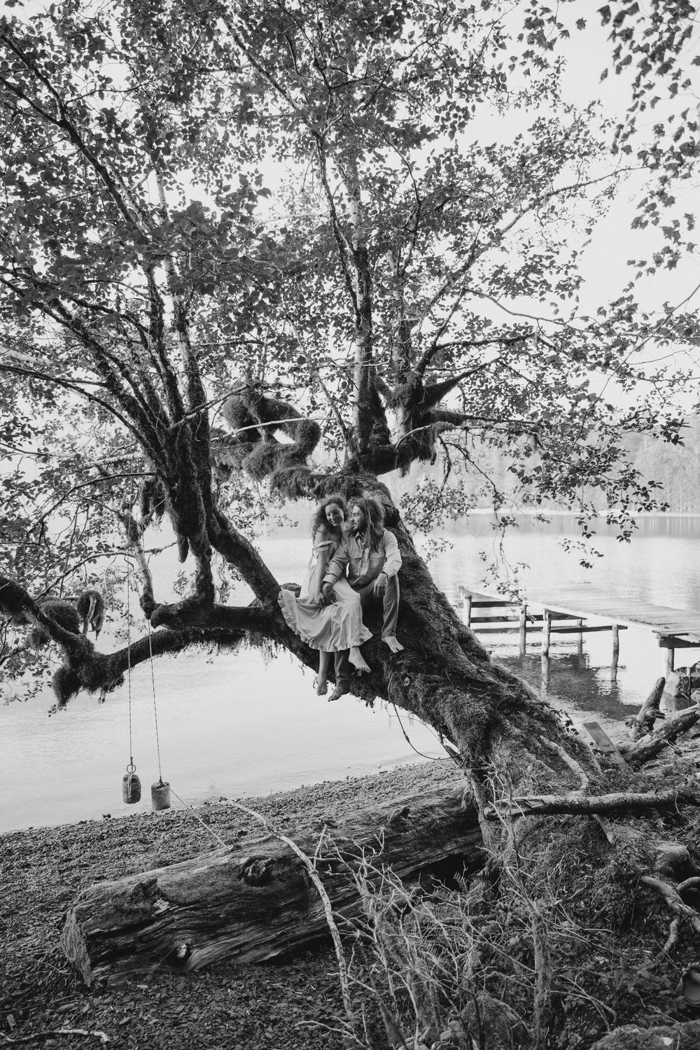Black and white photo of people sitting on a large tree branch overhanging a lake with a dock in the background.