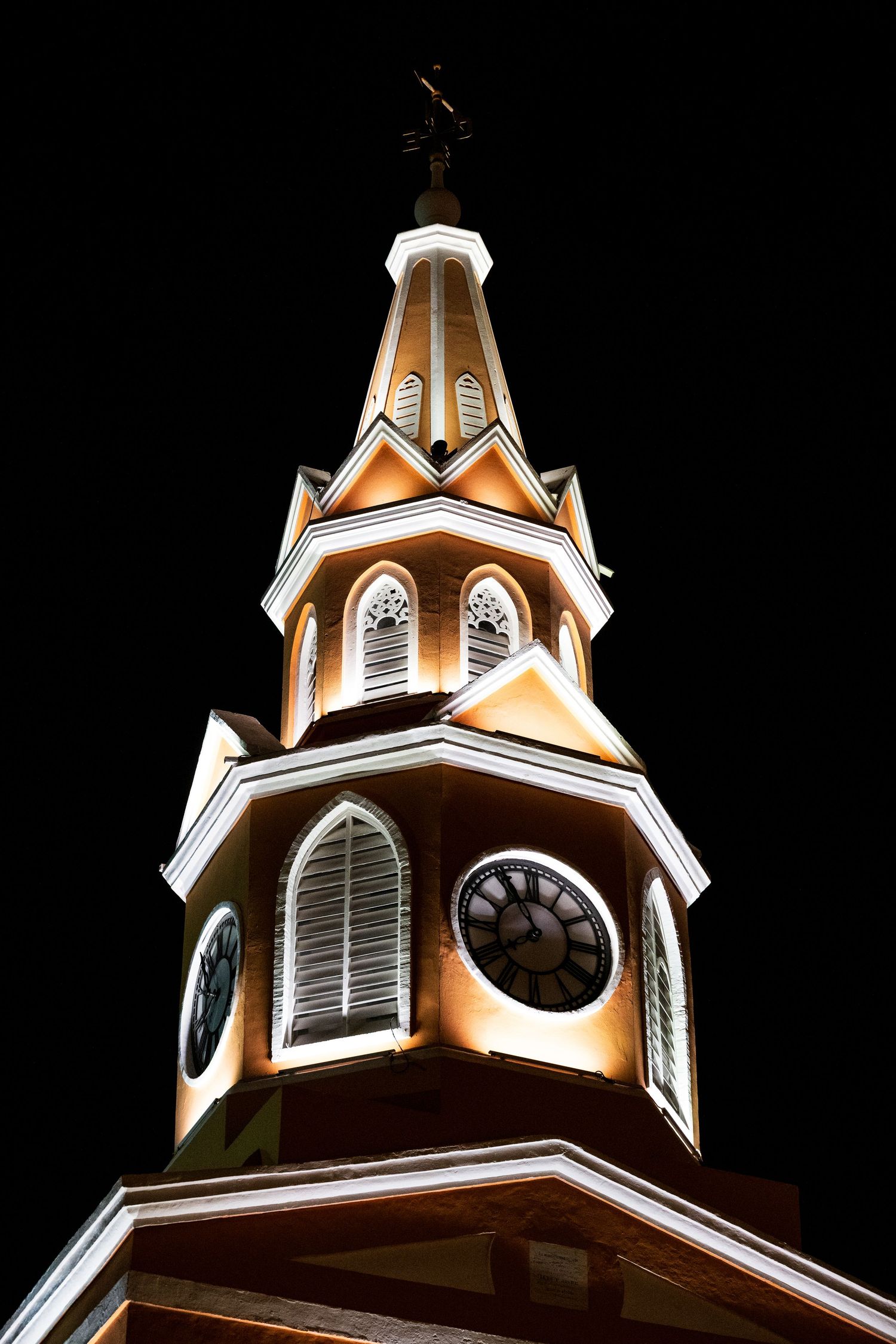 Illuminated church steeple with ornate white trim and clock faces against dark night sky.