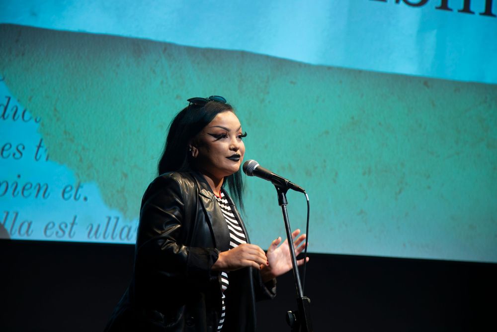A presenter speaks at a professional conference or event with blue lighting and projection screens in the background.