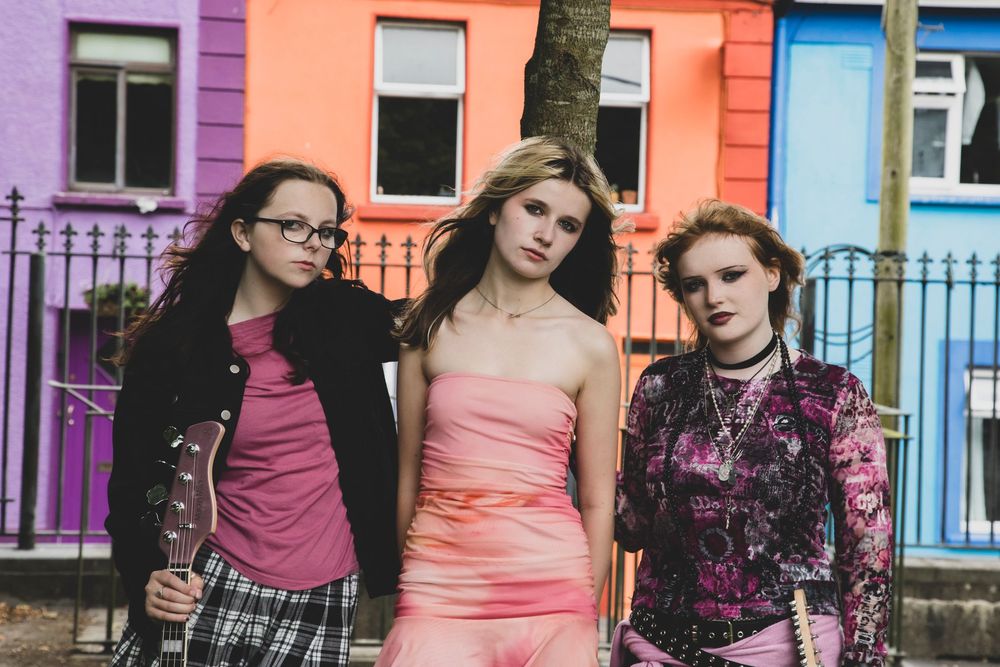 Three people pose together against colorful row houses wearing pink and black alternative fashion outfits.