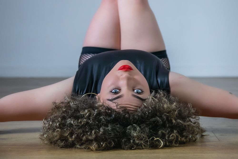 A person with curly hair and red lipstick lies upside down on a wooden floor wearing black clothing.