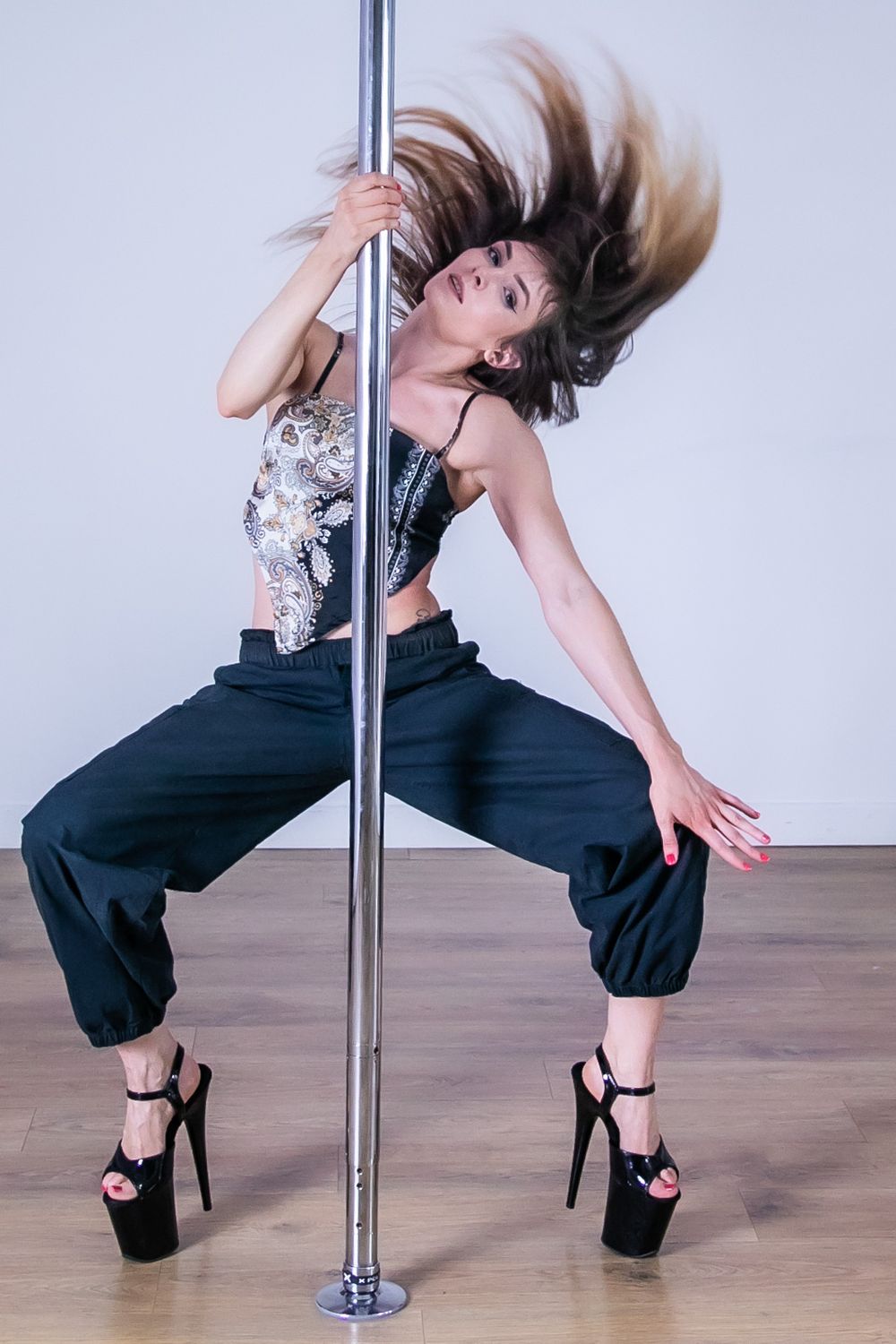 A pole dancer in black pants and silver top flips their hair while performing a dance move in platform heels.