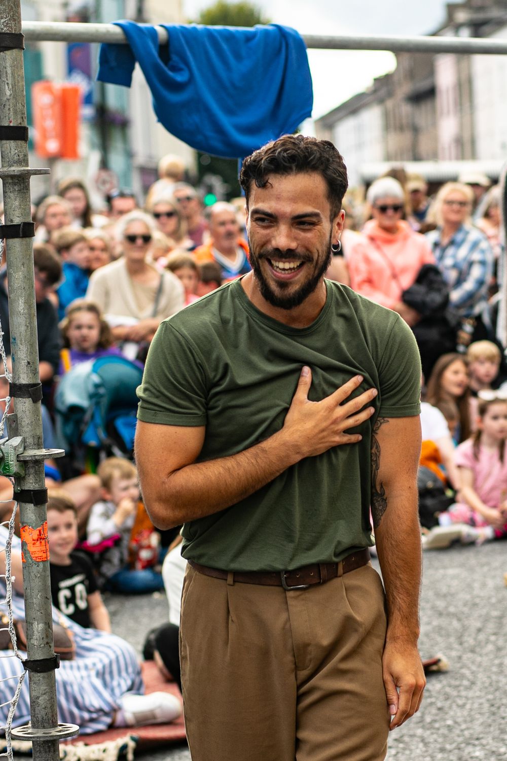 A performer in a green shirt laughs joyfully during an outdoor street performance with a crowd watching in the background.