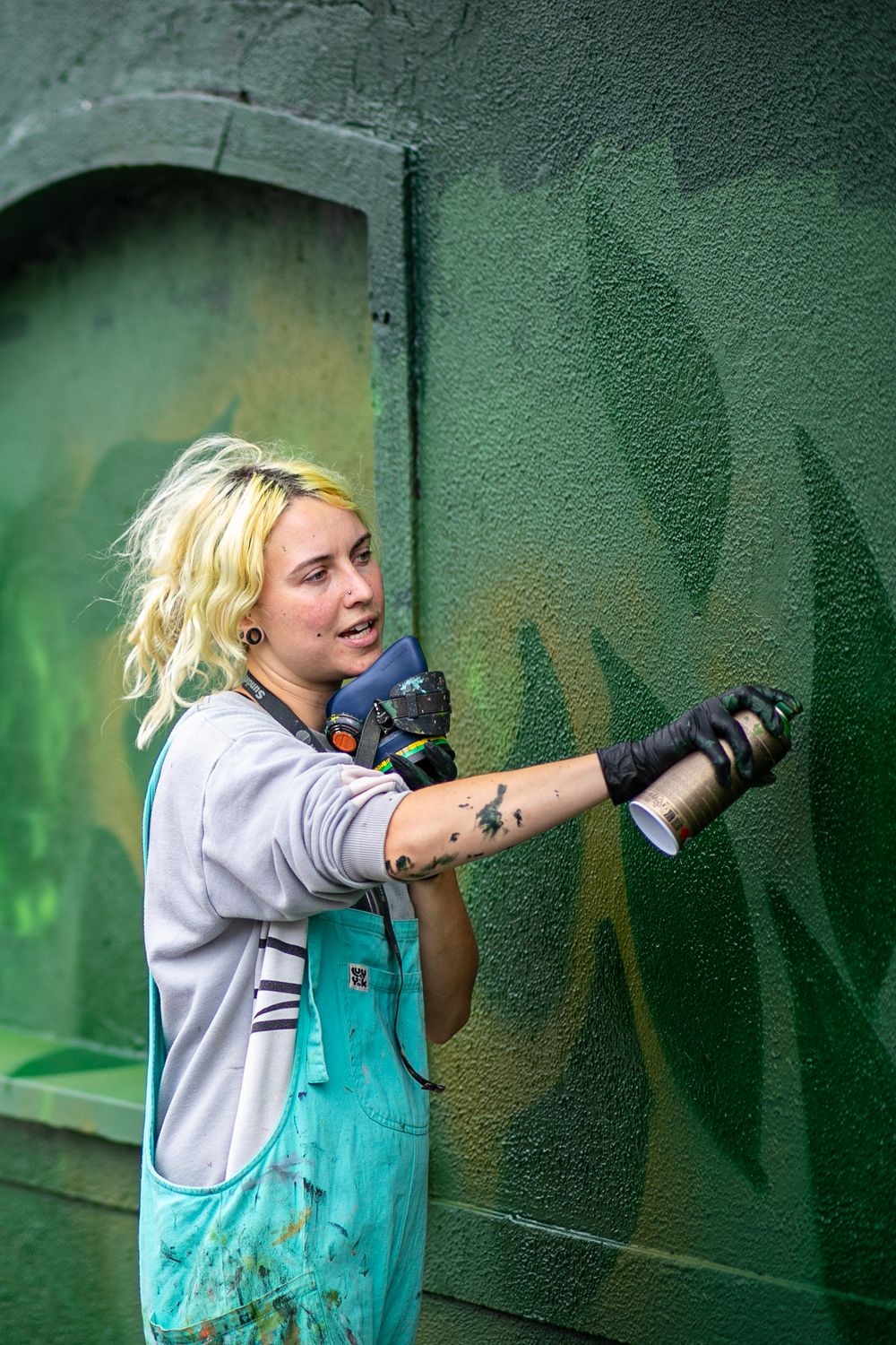 Artist in turquoise overalls works on painting a green wall.