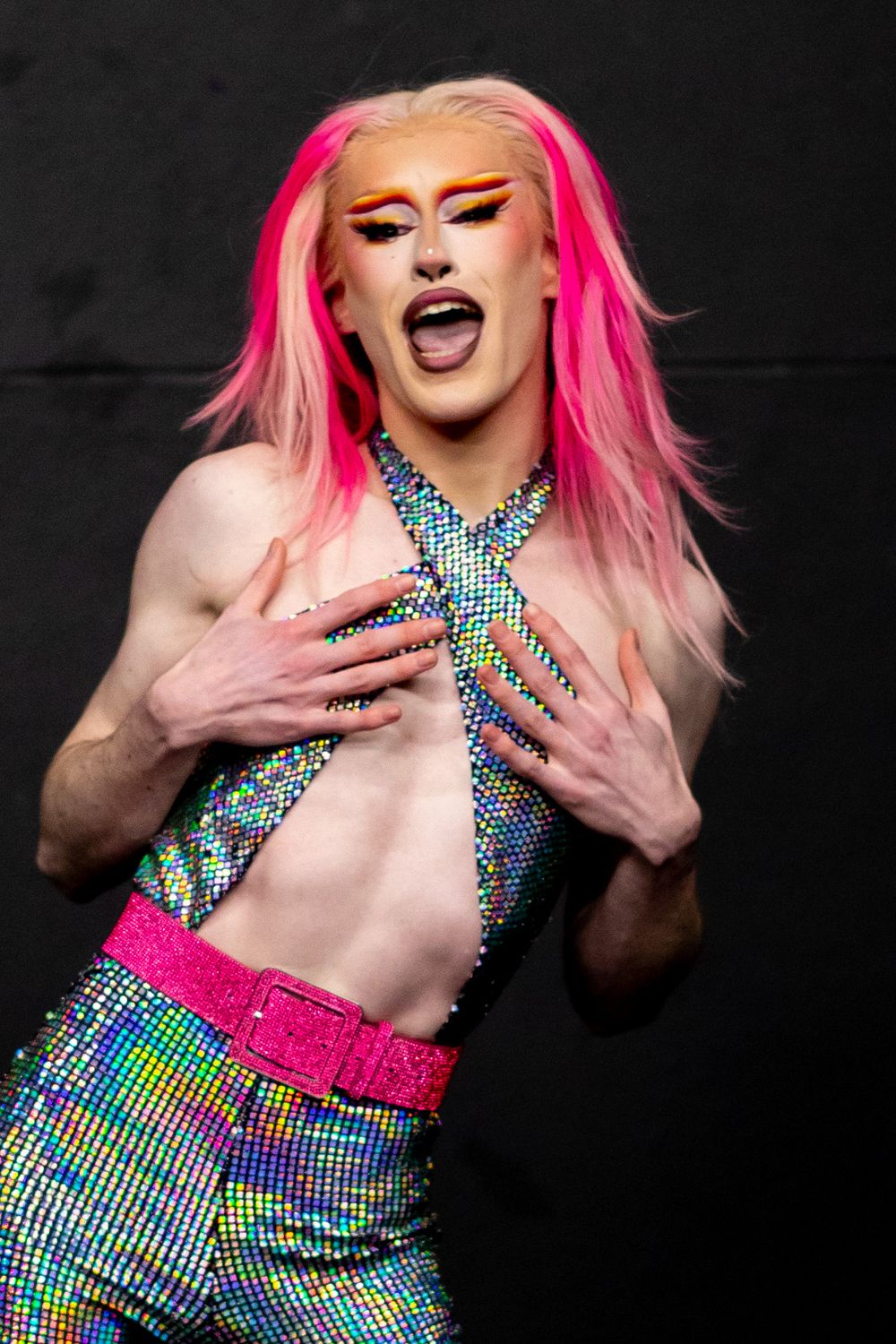 A performer in glittery holographic drag outfit with pink hair poses dramatically against dark background.