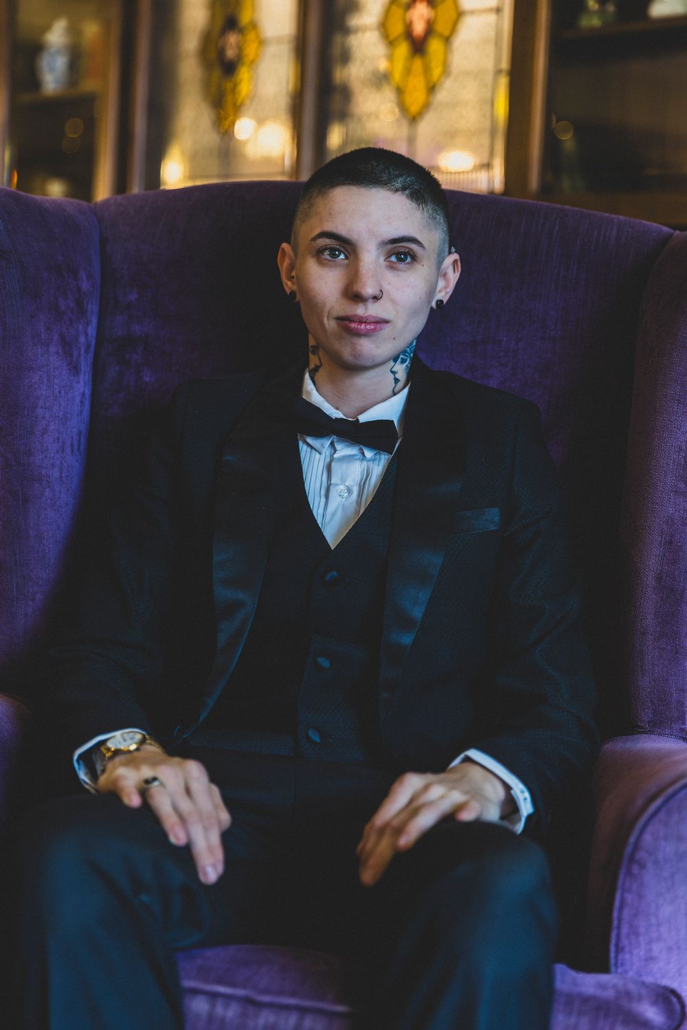 Young boy in formal black suit and tie sits on purple velvet chair in dark moody lighting.