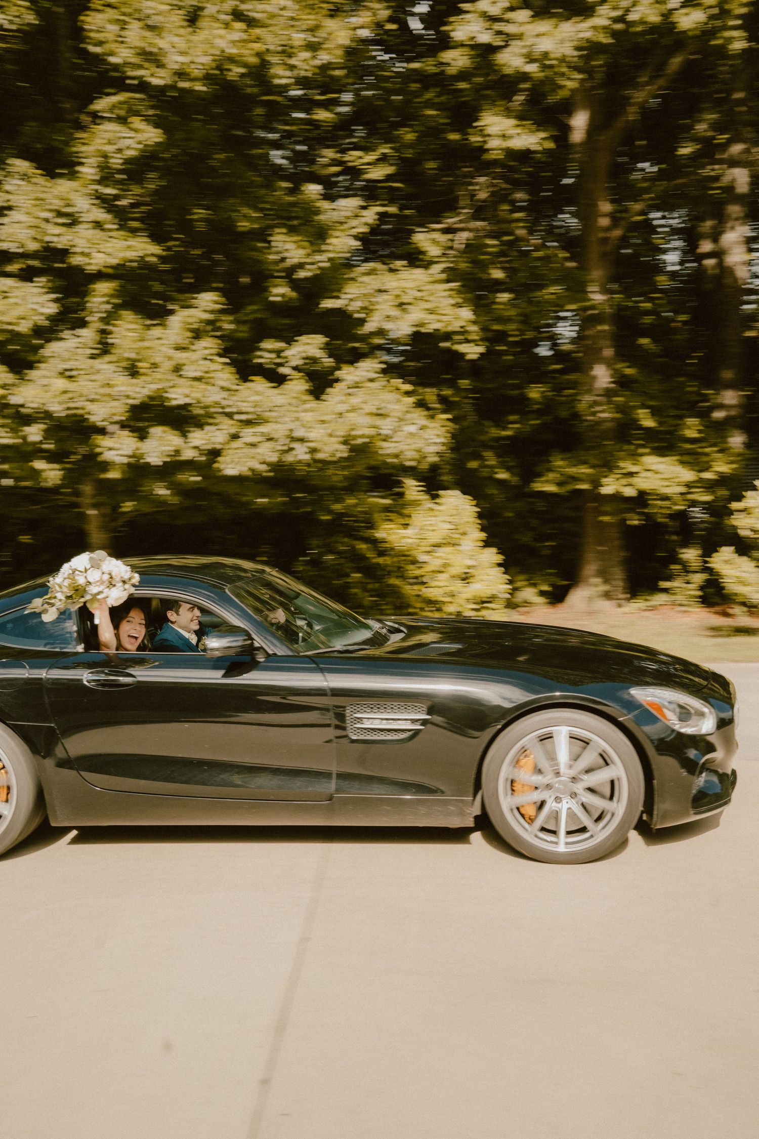 A black luxury Mercedes sports car drives on a tree-lined road with golden sunlight filtering through the foliage.