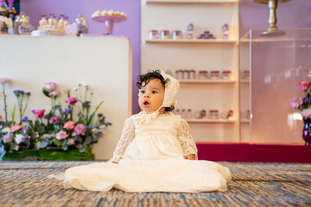 Baby in white lace dress sitting on textured floor with floral decorations in background.