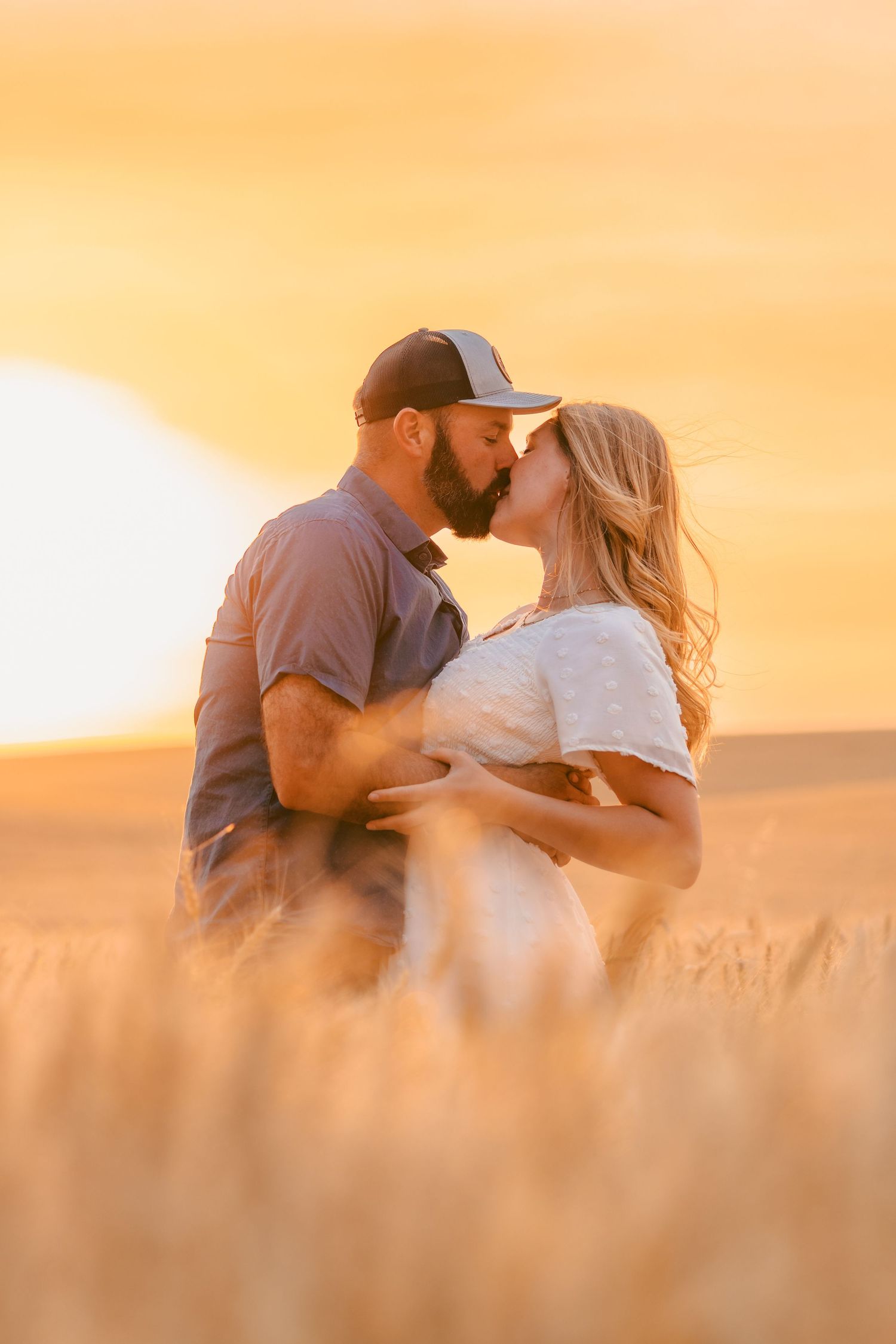 Couple kissing romantically in golden wheat field at sunset, man in cap embracing woman in white dress.