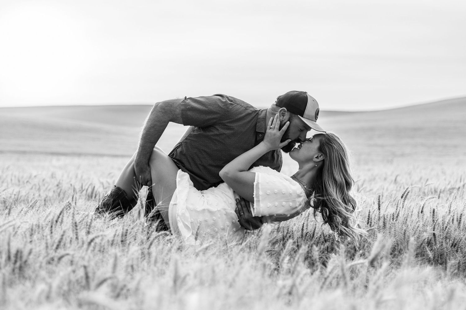 Black and white photo of a couple sharing a romantic dip kiss in a vast wheat field.