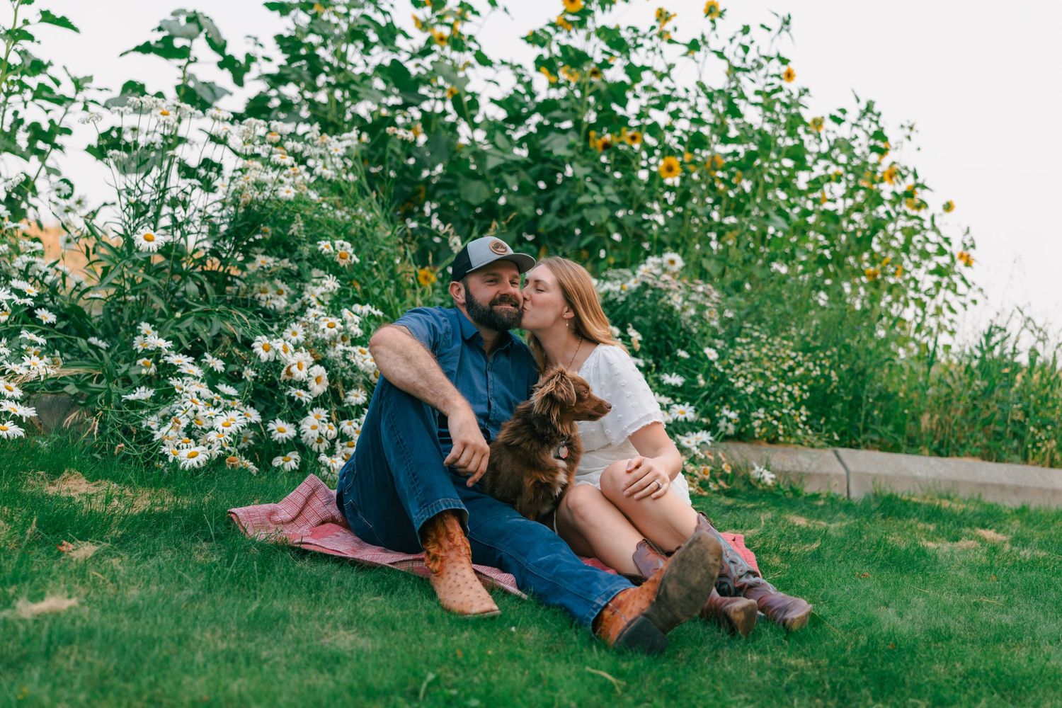 Couple sitting on grass with a dog, surrounded by blooming white flowers and sunflowers in a lush garden.