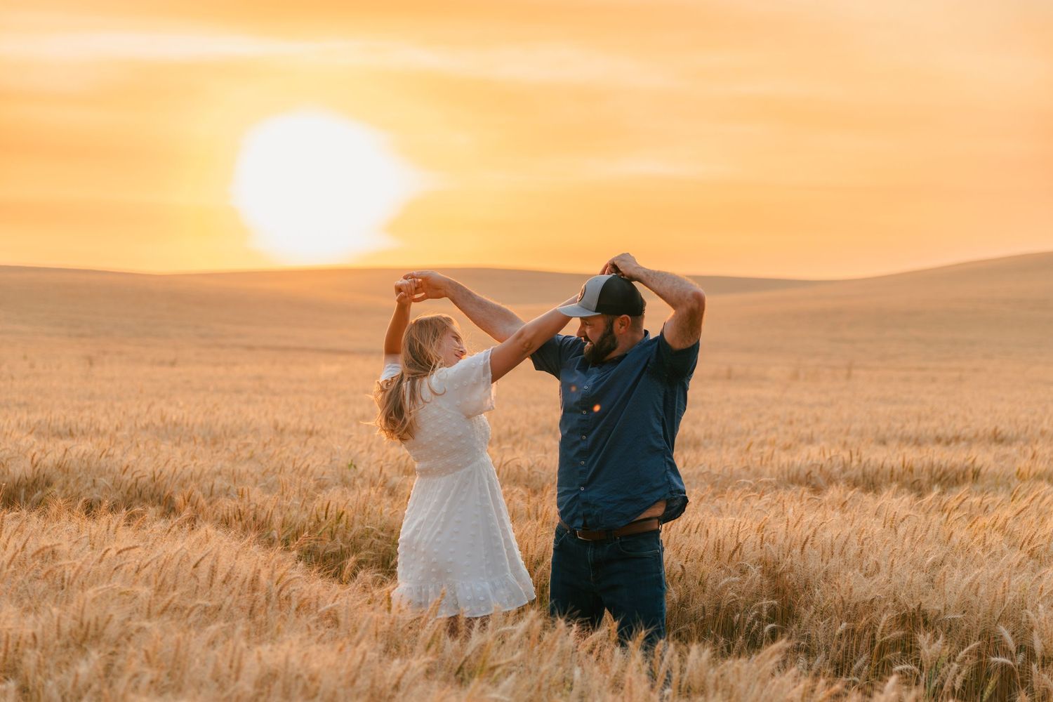 Couple dancing together in a wheat field beneath a vibrant golden sunset.