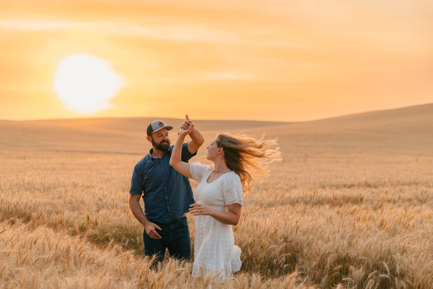 Couple dancing in golden wheat field at sunset, woman in white dress twirling under man's hand.