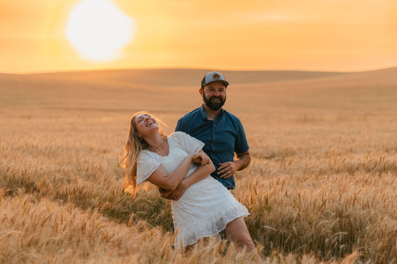 Joyful couple laughing and spinning in a golden wheat field at sunset.